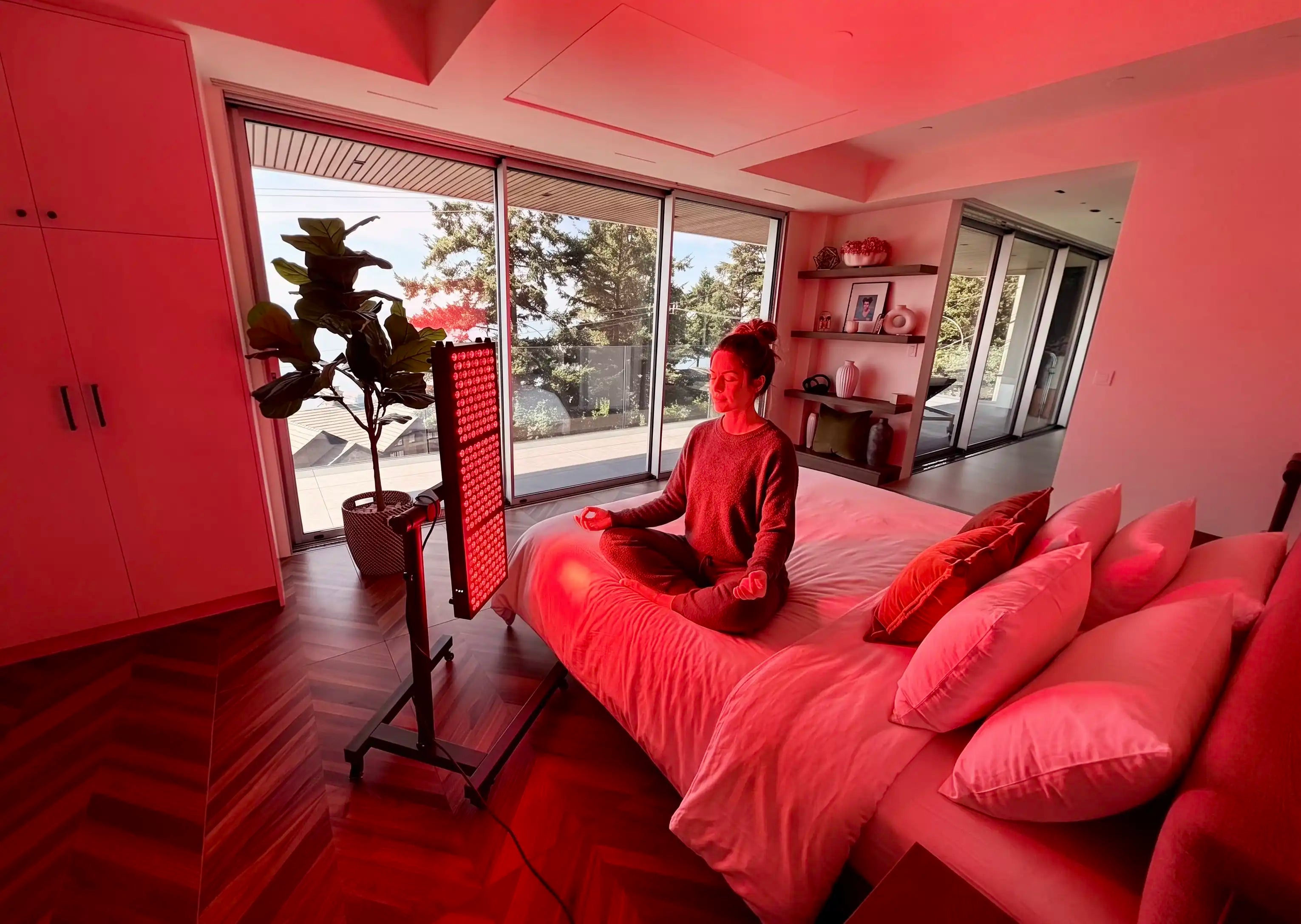 Person sitting on a bed in a modern room with large windows and red light therapy machine