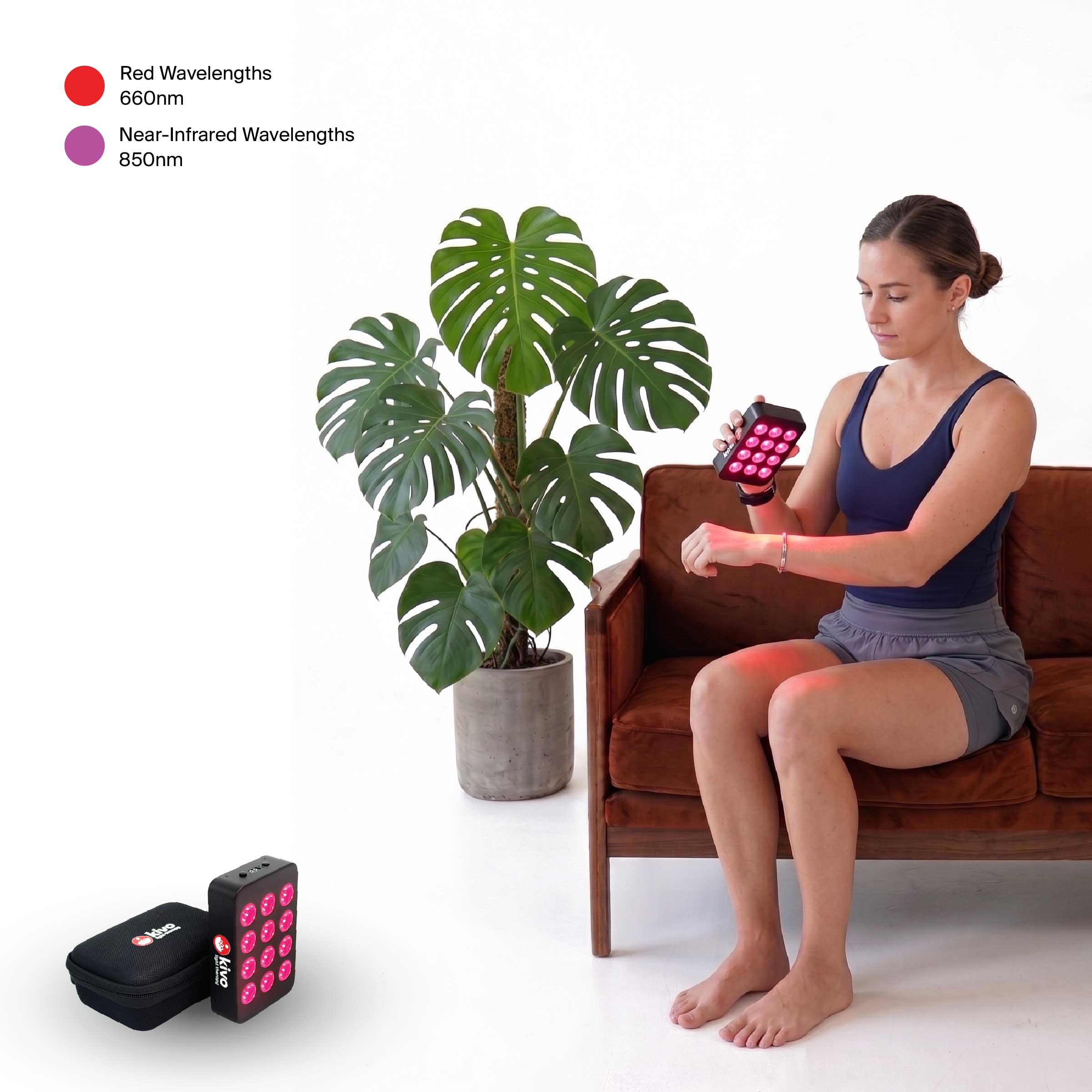 A woman sits on a brown leather couch using a Kivo Vital MINI red and near-infrared light therapy device