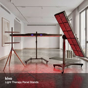 Three Kivo light therapy stands displayed in a modern, bright studio room with large windows and a concrete floor. The stands include a flat panel on a tripod base, a long horizontal panel on a central stand, and a large angled panel on a wheeled cart, all emitting red light.